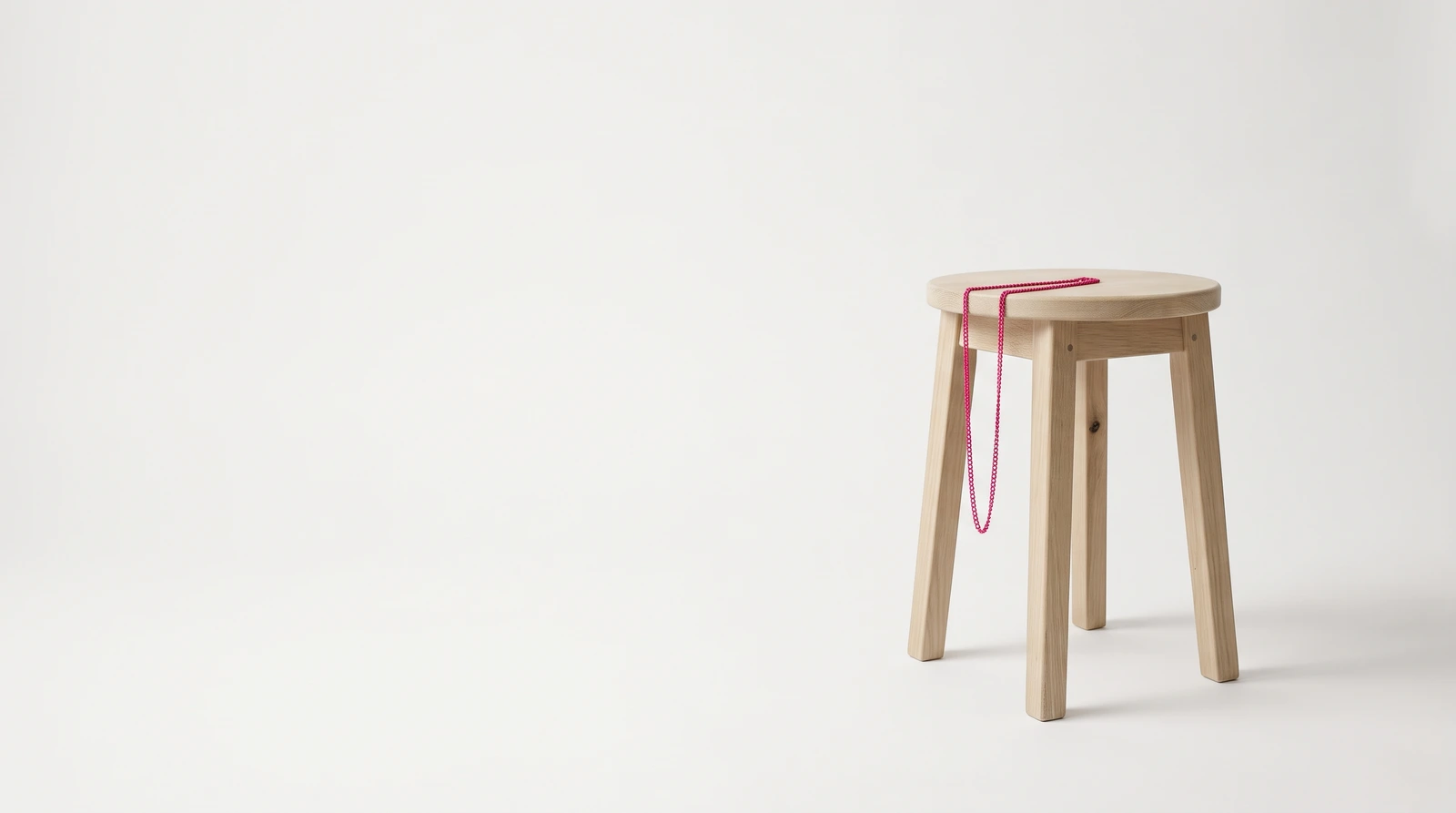 A single plain oak stool standing alone on an empty white studio sweep with nothing else in the room, a fine magenta chain draped over the near edge of its seat